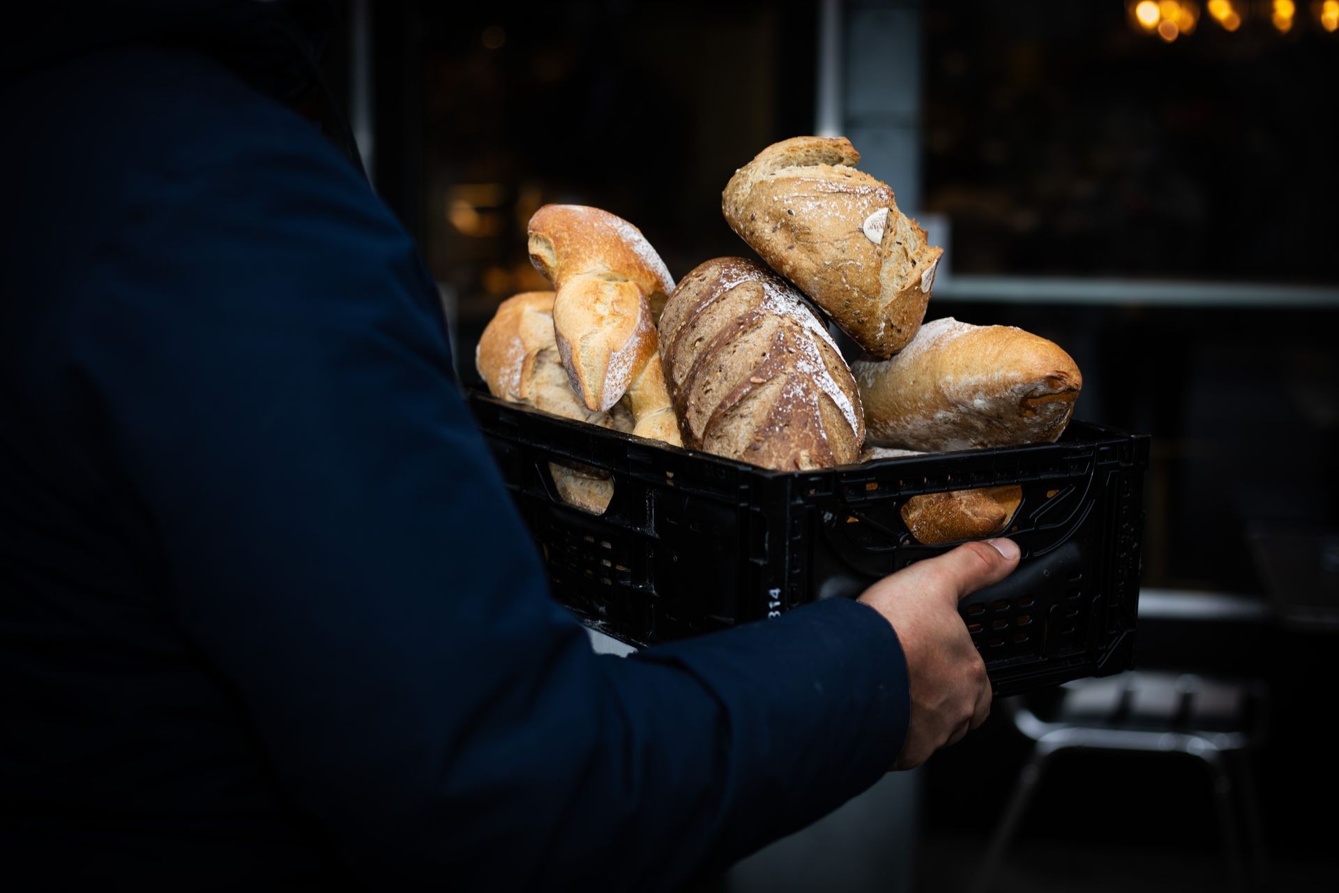 Person carrying a black crate filled with various loaves of freshly baked bread.